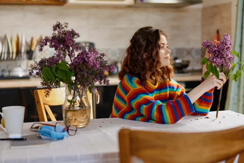 Young woman with curly hair sits at a table arranging a lilac bouquet in a bright kitchen, smiling softly, her striped rainbow sweater adding color as sunlight streams through the window for a warm,