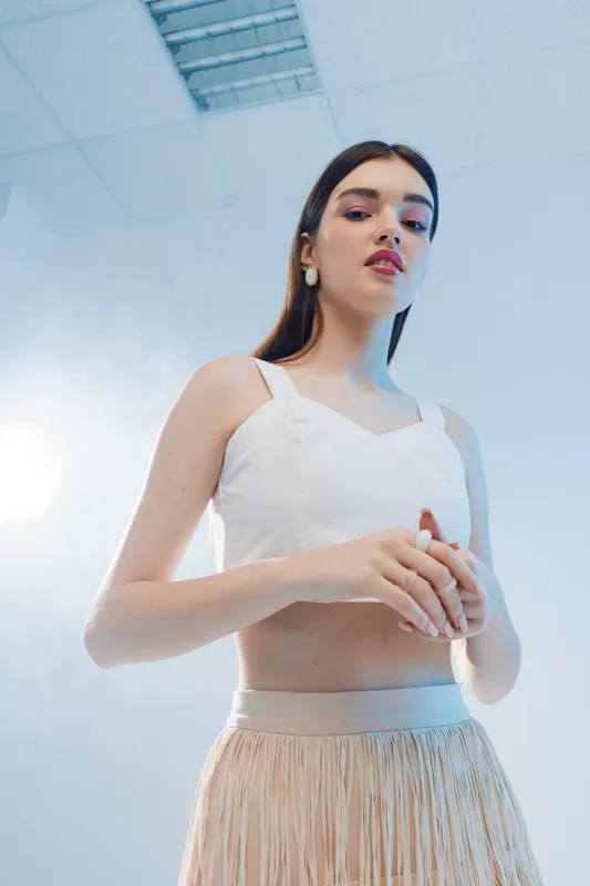 Young woman in white crop top and beige skirt poses confidently in a bright studio with modern ceiling lights. Concept of beauty, fashion, and contemporary style.