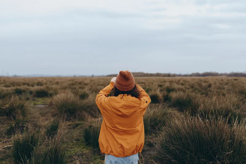 Woman in orange coat and knit hat, seen from back view, standing in an open grassy field outdoors, Winter nature landscape with calm lifestyle atmosphere, soft daylight, breeze moving grass