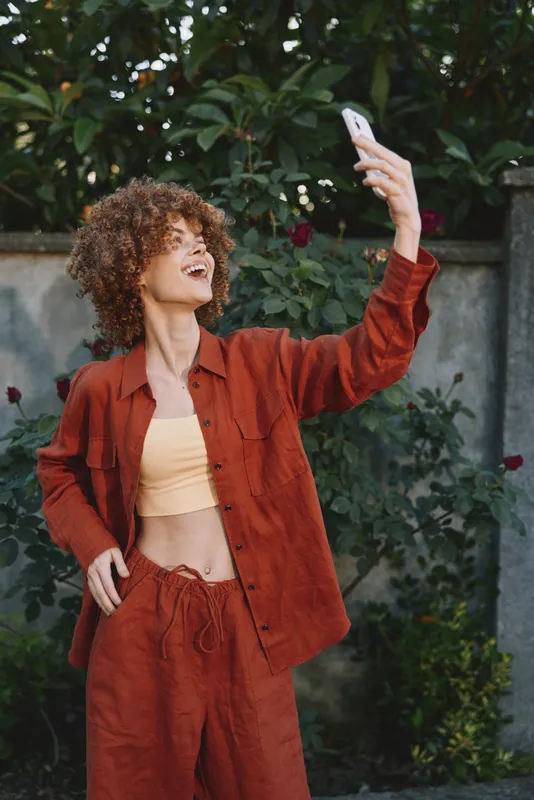 Smiling woman with curly hair takes a selfie in a garden, showcasing casual fashion and lifestyle. Warm natural light highlights greenery, rust tones, and vibrant texture