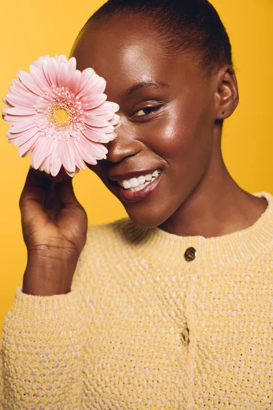 Smiling black woman with dark skin holds pink flower near eye, wearing yellow sweater. Studio portrait on yellow background. Concept of beauty, skincare, wellness, and natural happiness.