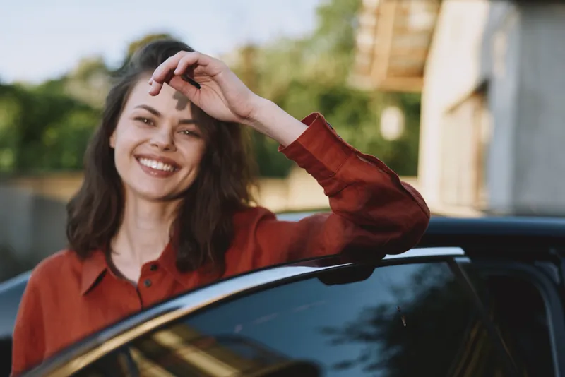 A smiling woman leans on a car in warm afternoon light, wearing a red shirt, relaxed pose. A lifestyle moment that conveys confidence, joy, and everyday happiness