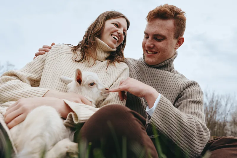Cheerful couple outdoors, smiling at a playful goat kid, cozy knitwear, carefree moment, natural colors, soft sunlight, relaxed posture, lush grass, gentle breeze, lifestyle scene, connection and joy