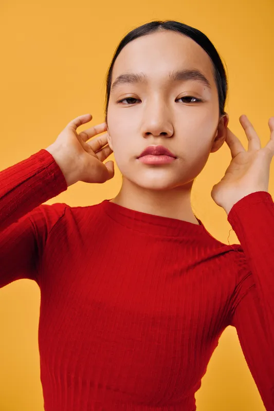 Young woman in red sweater posing with hands near ears on yellow background studio portrait. Asian female model with natural makeup and expressionless face for fashion concept.