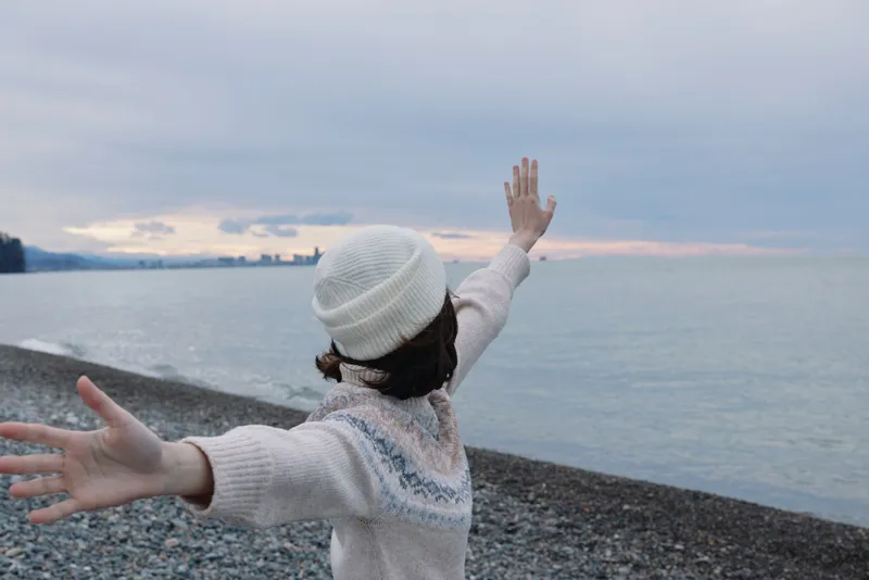 Woman with outstretched arms on a pebble beach by the sea at dusk, wearing a knit sweater and hat, expressing freedom, joy and calm in an outdoor winter scene.
