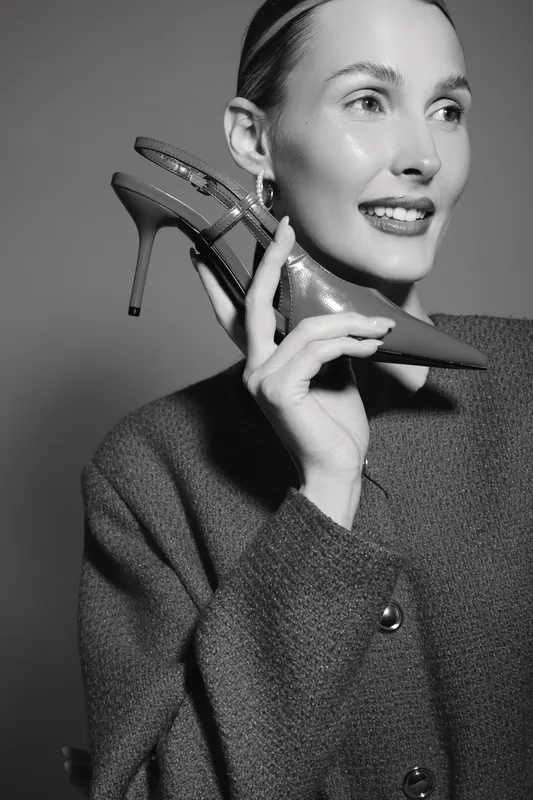 A smiling Caucasian woman with fair skin holds a high heel shoe near her face, wearing a textured coat. Concept for fashion, elegance, and style in studio lighting.