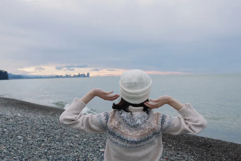 Beach, person, sea, back, sweater, hat scene of a person on a pebbled shoreline facing the horizon at dusk, a calm contemplative coastal moment.