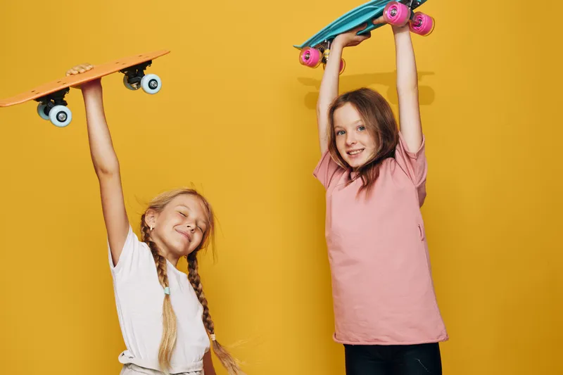 Two smiling girls holding colorful skateboards above their heads on a yellow background. They wear casual t-shirts and enjoy an active, playful moment together.