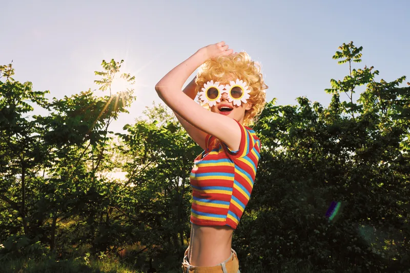 Playful child with sunflower sunglasses jumps outdoors in sunny park, wearing a striped rainbow top, capturing retro film color vibe and carefree summer mood, bright laughter, warm breeze, candid