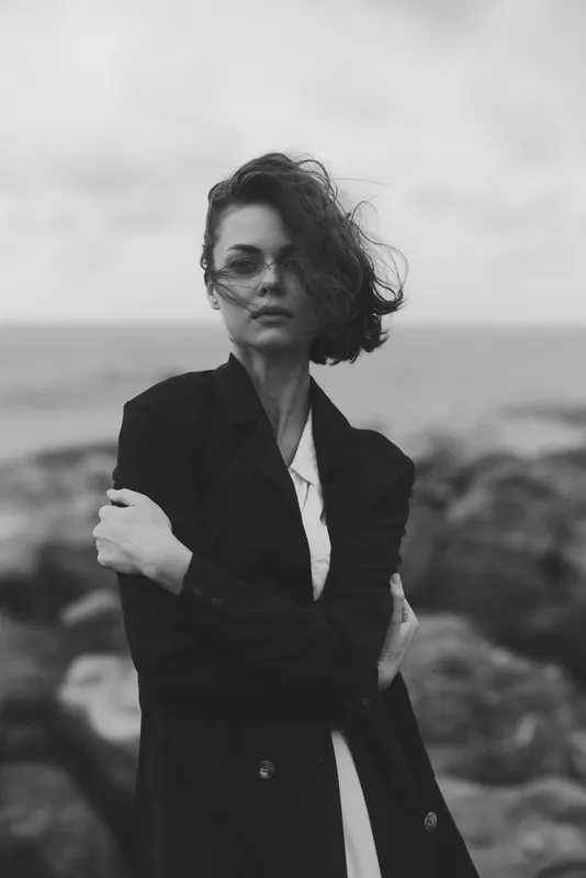 Woman in a tailored coat and sunglasses stands with crossed arms on a rocky shoreline, wind blowing hair, ocean haze in the background, monochrome mood, confident and stylish.