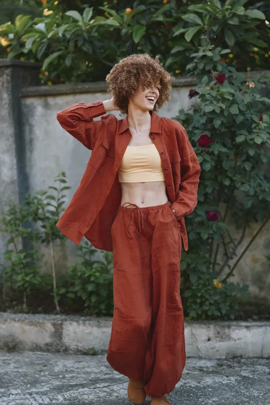 Young woman with curly hair wears rust suit and yellow crop top, posing outdoors with a cheerful smile, showcasing casual fashion and confident lifestyle in a relaxed garden setting with natural