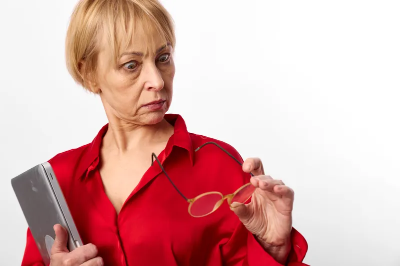 Elderly woman red shirt holding glasses and smartphone, concerned portrait on white background, indoor close up, searching focus, copy space.