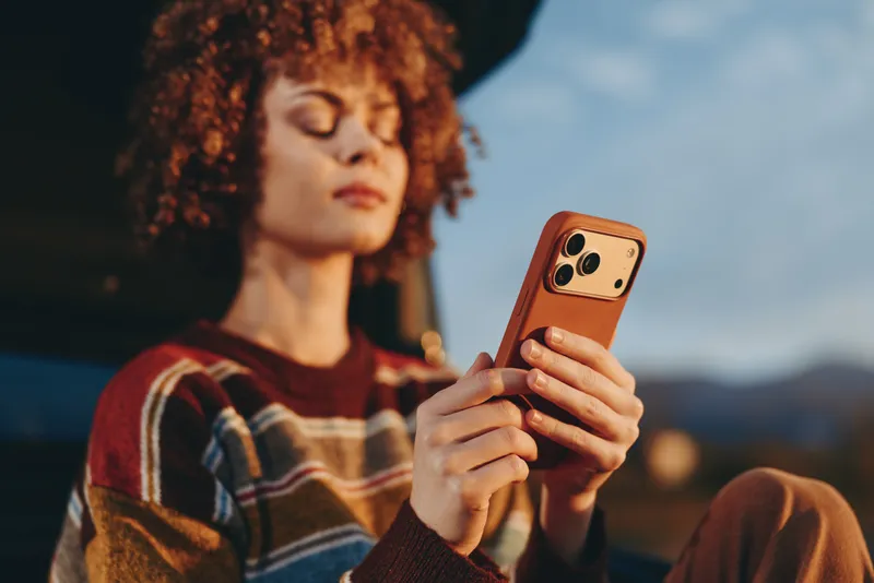 Woman lifestyle outdoors, curly haired woman holding smartphone at golden hour, eyes closed, relaxed posture, natural light, soft breeze, mountains blurred behind, evening warm colors