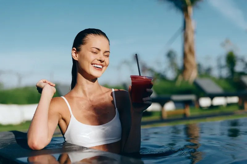Joyful woman in a white swimsuit relaxes at pool, under bright sun, holding a refreshing drink, exuding summer leisure and vacation vibes.