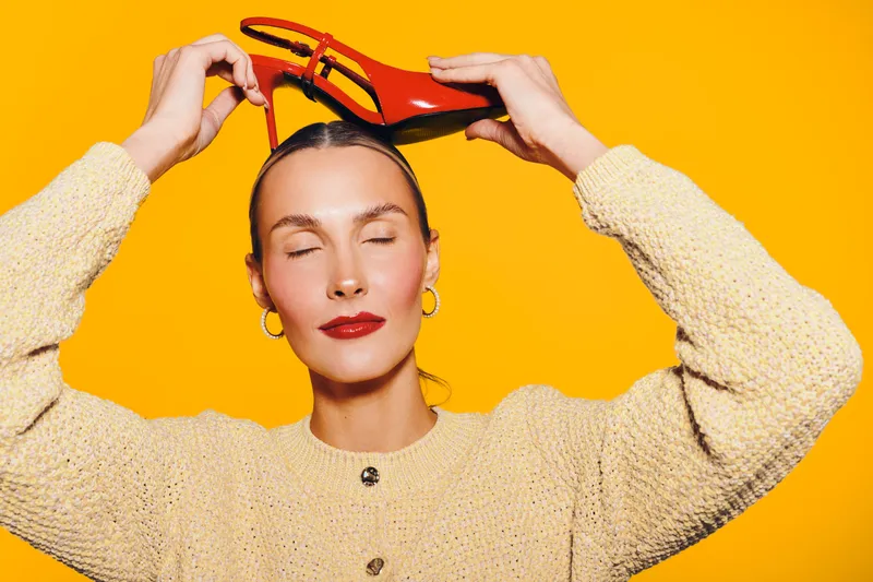 Young woman with closed eyes holds red high heel shoe above head on bright yellow background. Concept of fashion, style, beauty, and creative portrait with vibrant colors and elegance.