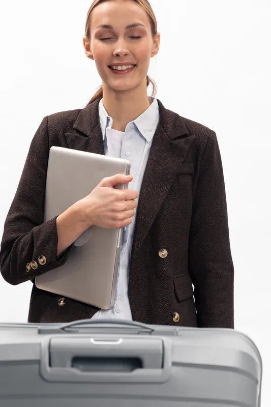 Smiling businesswoman in blazer holding laptop stands behind suitcase on white background. Concept of business travel, professionalism, and work preparation for trip or meeting.
