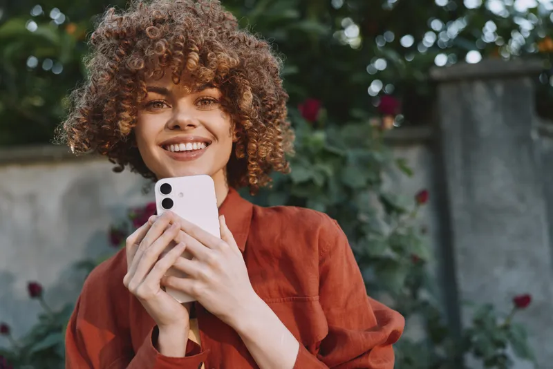 Young woman with curly hair using a smartphone outdoors, smiling in a garden setting, casual lifestyle moment, natural daylight, candid portrait and relaxed mood