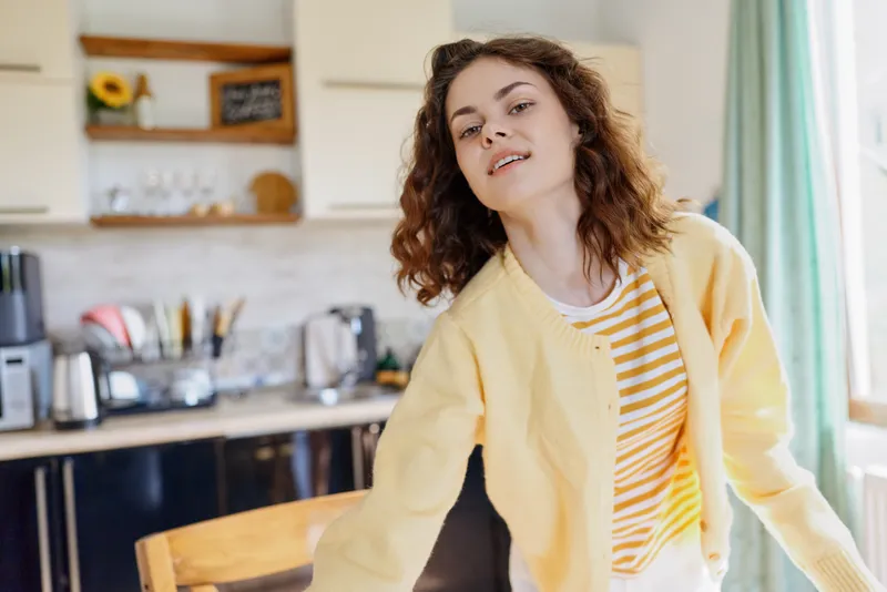 A cheerful woman stands in a bright kitchen, leaning on a chair, wearing a yellow cardigan and striped shirt. A warm, casual lifestyle moment at home.