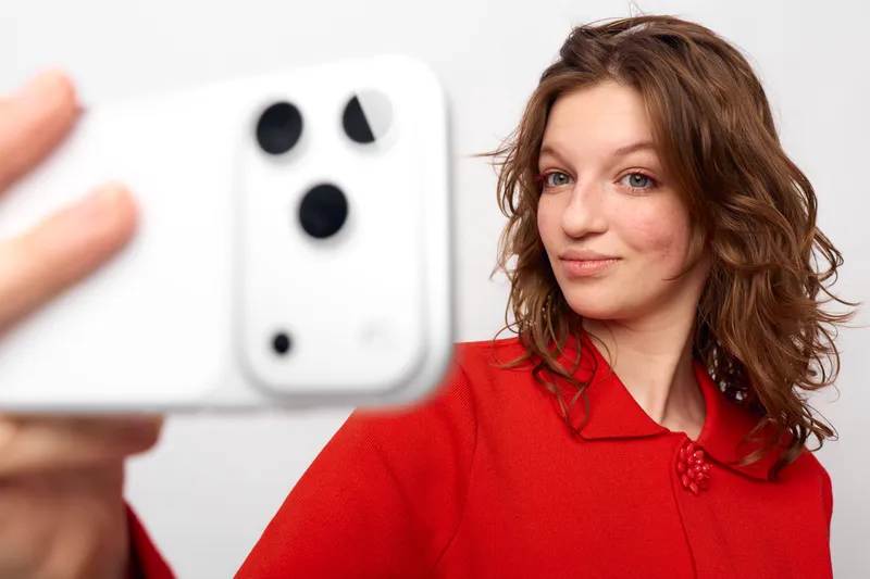 Young woman with curly hair wearing red sweater takes selfie with white smartphone on white background. Concept of casual lifestyle, social media, and modern technology use.