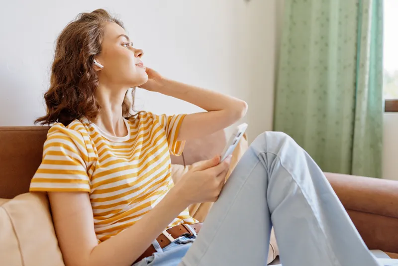 A woman relaxes on a sofa at home, wearing a striped t shirt and jeans, looking out the window with a thoughtful smile while holding a smartphone. Cozy, casual lifestyle image.