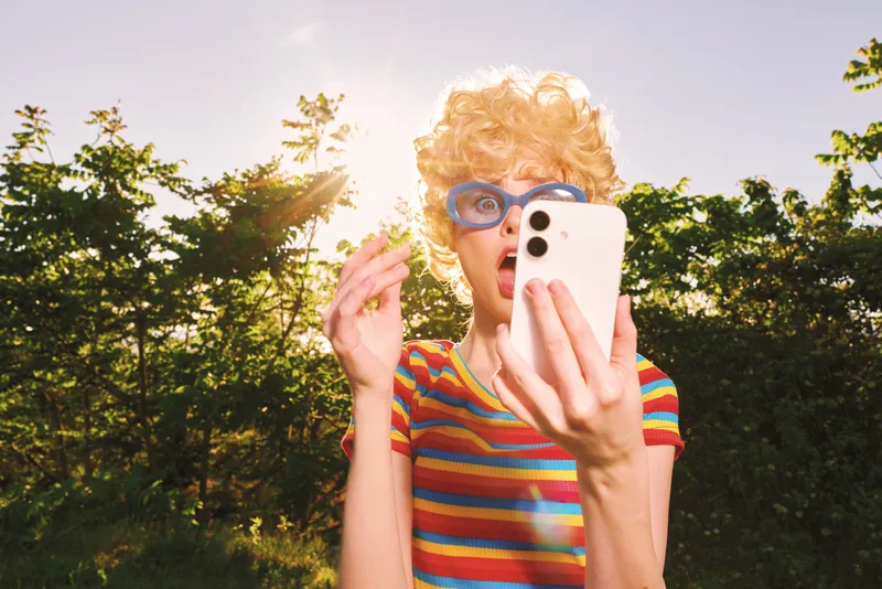 A playful woman outdoors wears a striped shirt and blonde wig, holding a smartphone for a selfie. Bright sun and trees create a warm retro film vibe.