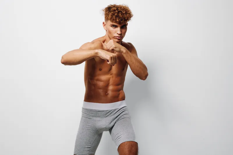 Young fit man training and posing as boxer with barechest, showing muscles and abs, wearing sports shorts, isolated on white background with intense focused expression.