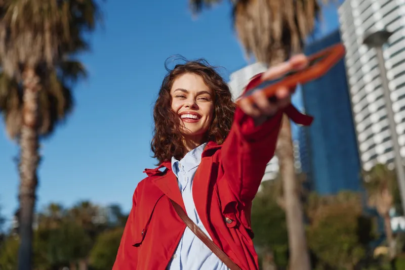 Smiling young woman in red jacket holds out smartphone taking selfie outdoors with palm trees and blue sky background, enjoying sunny day in urban environment.