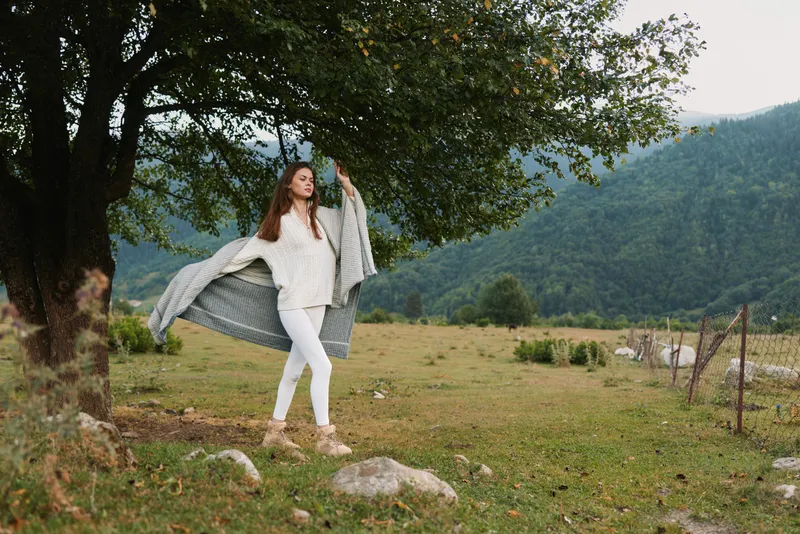 Young woman outdoors walking near tree in open field with mountains background wearing white clothes and gray shawl, enjoying nature and fresh air, casual lifestyle.