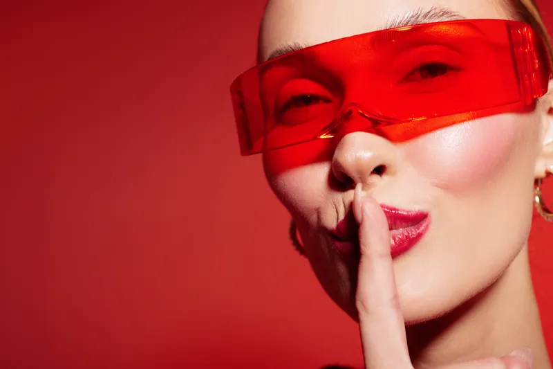 Close-up of a woman wearing red transparent glasses making a shushing gesture with finger on lips. Red background creates bold and mysterious mood. Concept of secrecy and silence.
