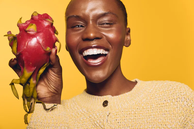 Happy African woman with dark skin holding a ripe dragon fruit on yellow background. Close-up portrait with bright smile and natural texture. Concept of healthy food and joy.