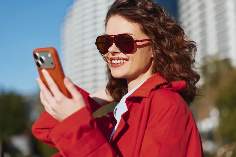 Young woman in a red jacket outdoors, wearing sunglasses, smiling at her phone as sunlight bathes an urban park, capturing a candid friendly moment for social media