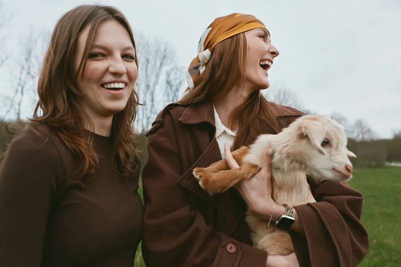 Woman, women, goat, kid, outdoors, happiness, friendship, lifestyle, animal, field, Two joyful friends pose outside with a playful goat kid, wearing warm jackets, capturing a candid outdoor moment.