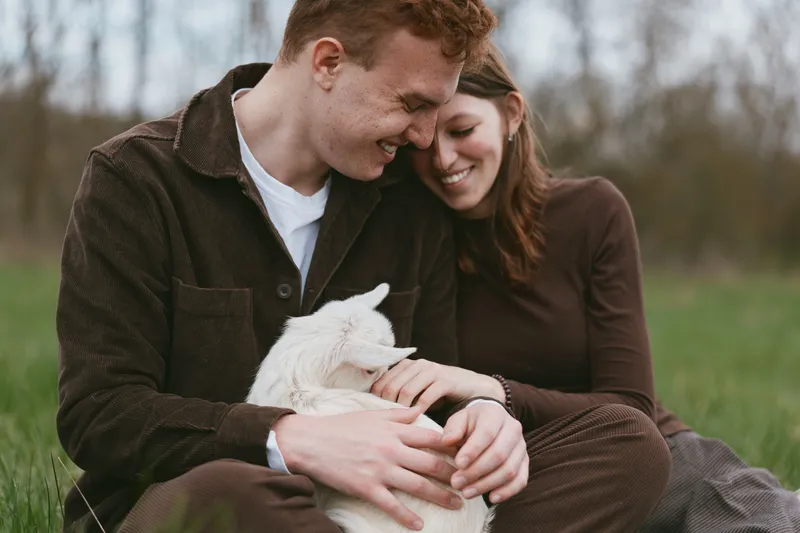 Cheerful couple outdoors, cuddling a white goat kid, warm sunlight, relaxed posture, gentle touch, grassy meadow backdrop, candid moment, natural colors, simple joy, lifestyle scene