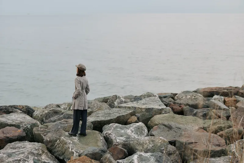 A woman in coat and hat stands on large rocks facing calm sea under overcast sky. Concept of solitude, contemplation, and peaceful coastal landscape in cool muted colors.