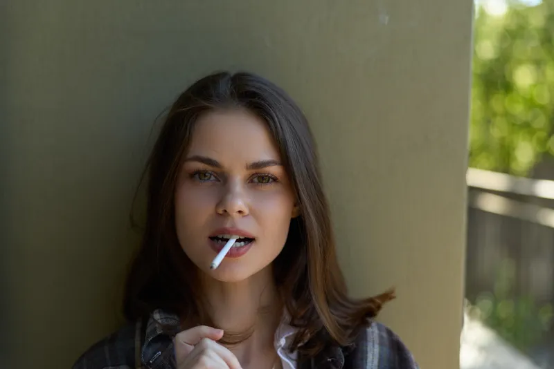 Casual woman smokes cigarette near a pillar outdoors, relaxed lifestyle portrait with urban park vibe, thoughtful and calm mood, natural light