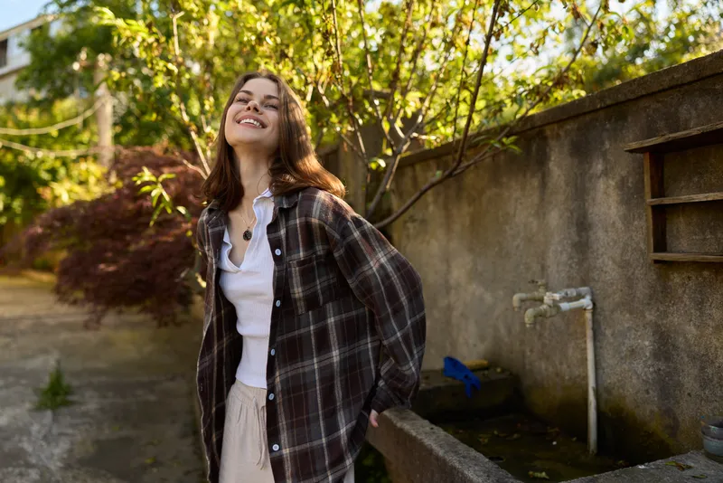 Bright portrait of a smiling woman outdoors in a casual plaid shirt, sunshine lighting, relaxed mood in a garden setting, natural background, everyday fashion and candid confidence