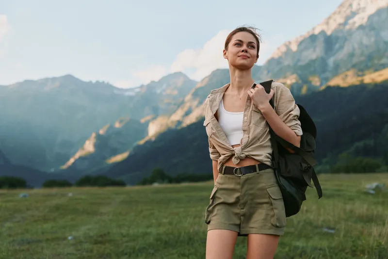 Woman adventurer stands in a mountain meadow, wearing a tied shirt and shorts, with a backpack, enjoying nature and open landscape under blue skies with distant peaks.