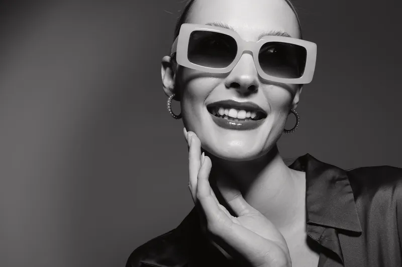 Happy Caucasian woman with pale skin wearing large sunglasses and hoop earrings smiles confidently in studio. Concept of fashion, style, and beauty portrait.