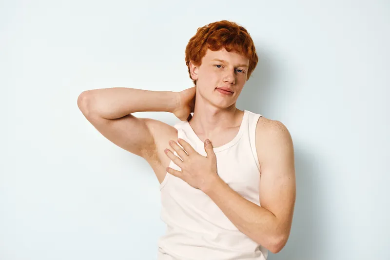 Young man with red hair in white tank top touching his neck and chest standing against white background with relaxed and confident expression in casual pose.