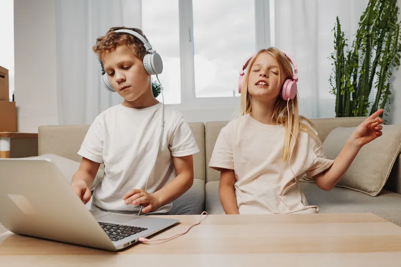 Two children wearing headphones enjoy music at home, one using laptop while the other sings with eyes closed, cozy living room setting.