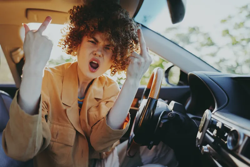 Woman with curly hair sits inside a car, making rock hand gestures, rebellious expression, retro film color palette, daylight through window, casual lifestyle portrait