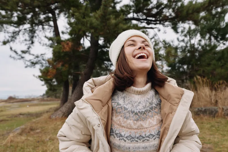 Woman outdoors, smiling, wearing a warm jacket, knit sweater and beanie, enjoying fresh air in a natural park setting with trees and open space, bright mood and carefree atmosphere.