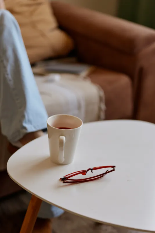 Close up of a white mug with tea on a round table, eyeglasses nearby, in a cozy living room with a soft blanket and sofa, conveying home lifestyle, calm routine, and quiet relaxation.
