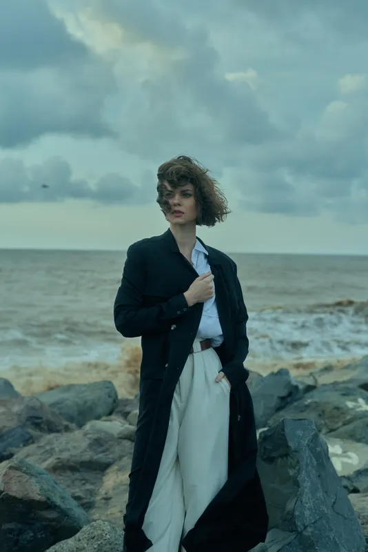 Woman in a long dark coat and light pants stands on a rocky beach, wind-swept hair, calm sea in the background, moody skies, fashion inspired, outdoor shoot, elegant and contemplative