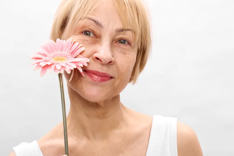 Older woman portrait, with a gentle smile while holding a pink gerbera flower by her face on a clean white background, beauty and calm, soft focus, in spring.