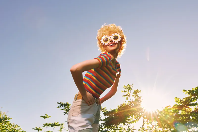 Young woman in a bright striped crop top and white shorts poses playfully outdoors with oversized flower sunglasses under a sunny blue sky, embodying carefree summer lifestyle.