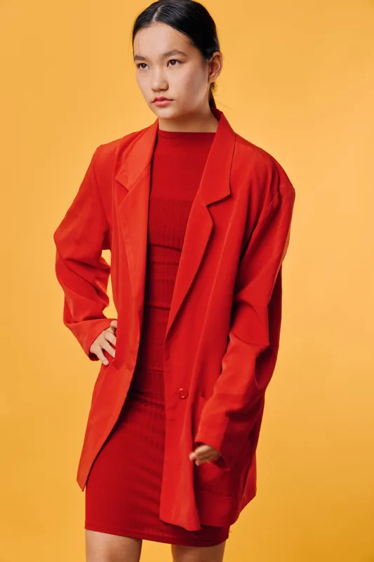 Woman wearing red oversized blazer and matching dress posing with hand on hip against yellow background. Fashion model with sleek hairstyle and confident expression in studio lighting.