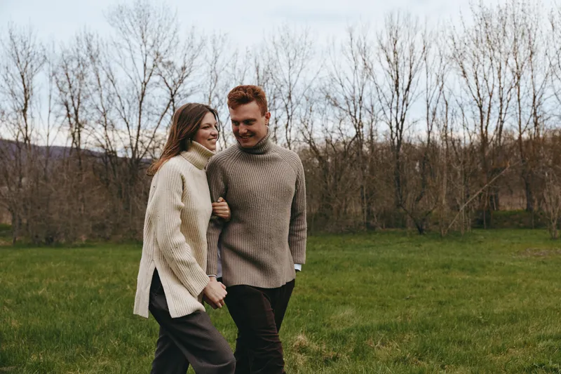 Smiling couple walking in a field wearing cozy knit sweaters. Casual outdoor moment highlighting connection and comfort. Soft light and relaxed mood convey wholesome lifestyle.