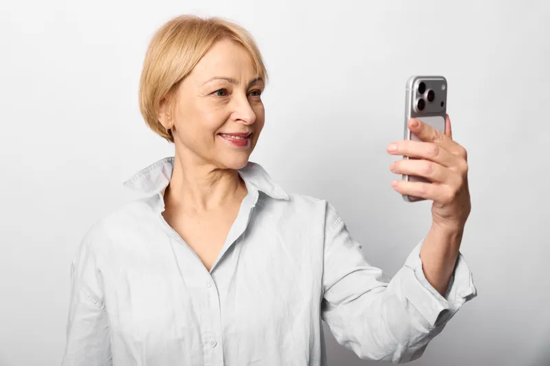 Smiling elderly woman holding smartphone for a selfie, studio portrait on white background, relaxed posture, soft lighting, minimal look, with gentle smile.