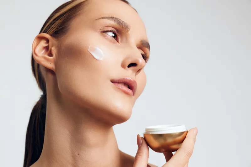 Close-up of young woman applying white face cream on cheek holding golden jar. Skincare and beauty concept with natural makeup and soft lighting on white background.